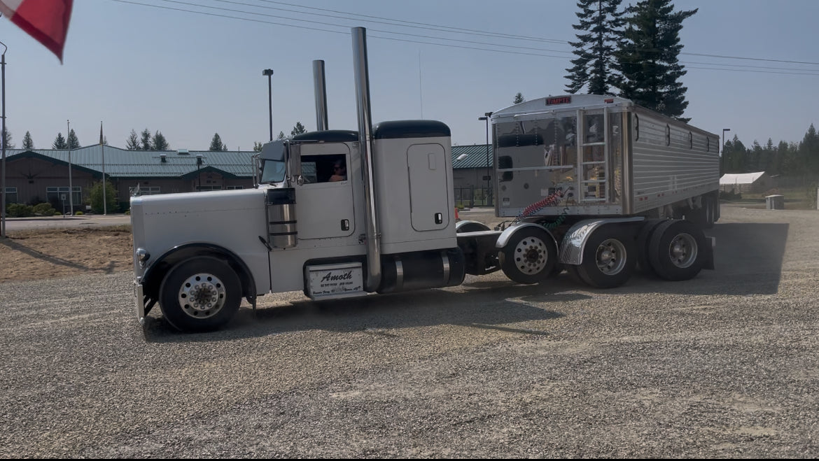 Truck with a trailer on a gravel lot with buildings and trees in the background